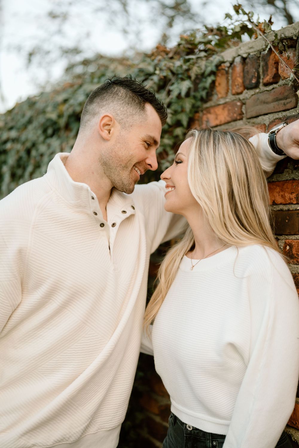A romantic couple shares a joyful moment leaning against a brick wall with ivy, wearing light cream and white sweaters.