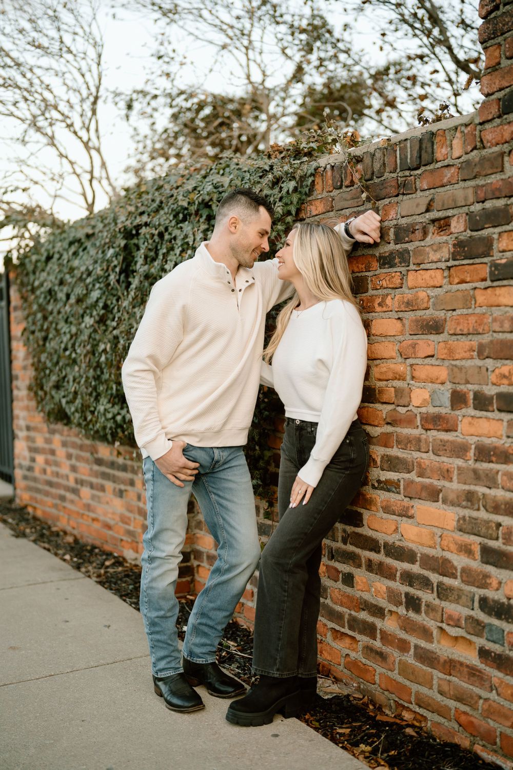 A romantic couple in casual attire leans against a rustic brick wall with ivy growing in the background.