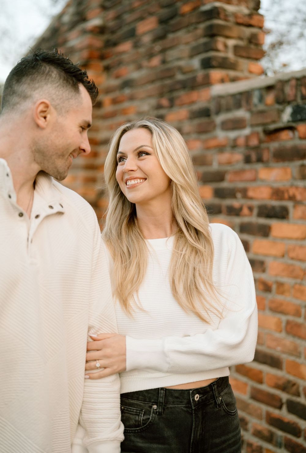 A loving couple shares a tender moment against a rustic brick wall backdrop, both wearing cozy cream sweaters.