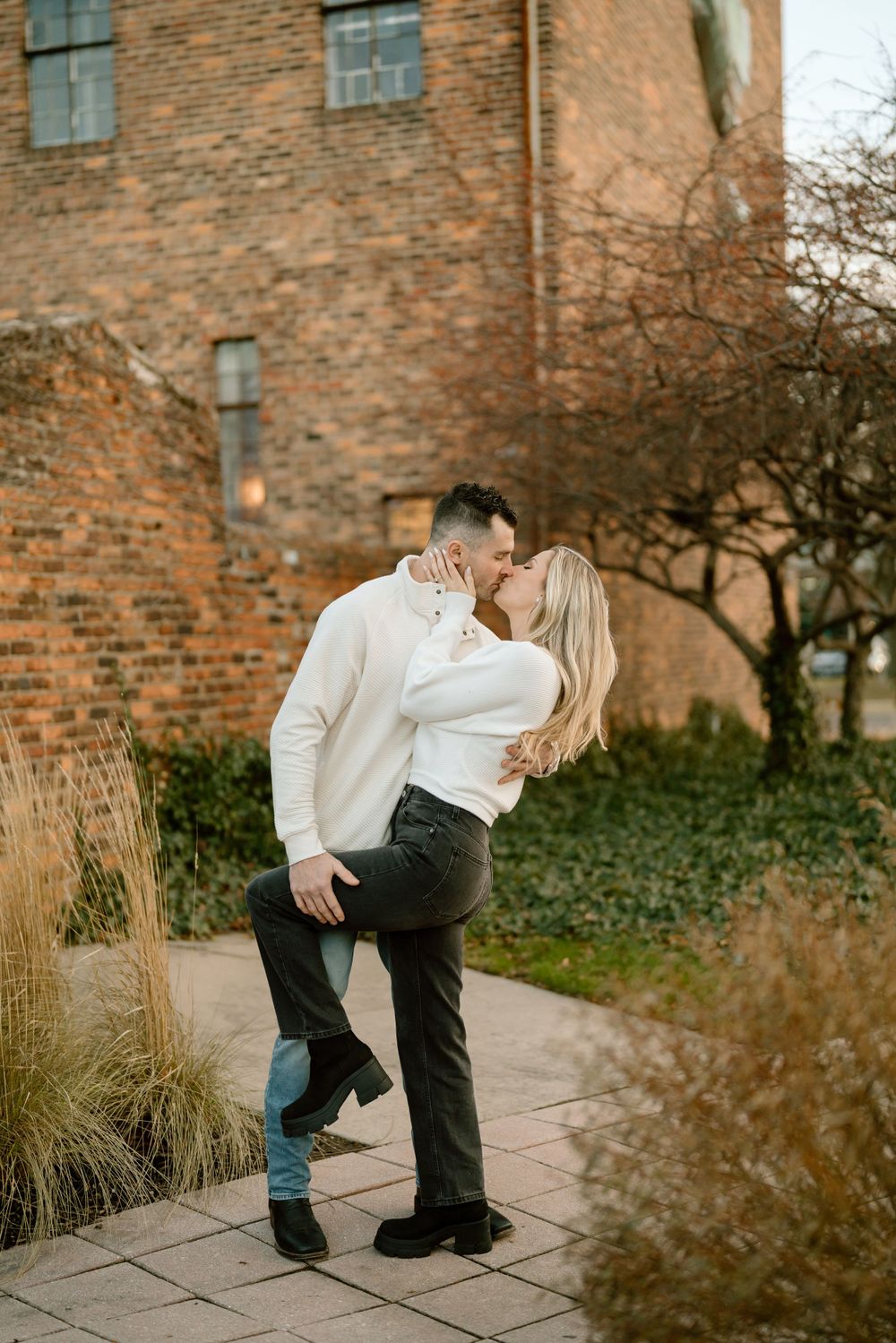 Romantic couple embracing by brick building with ornamental grasses and garden path in autumn setting.