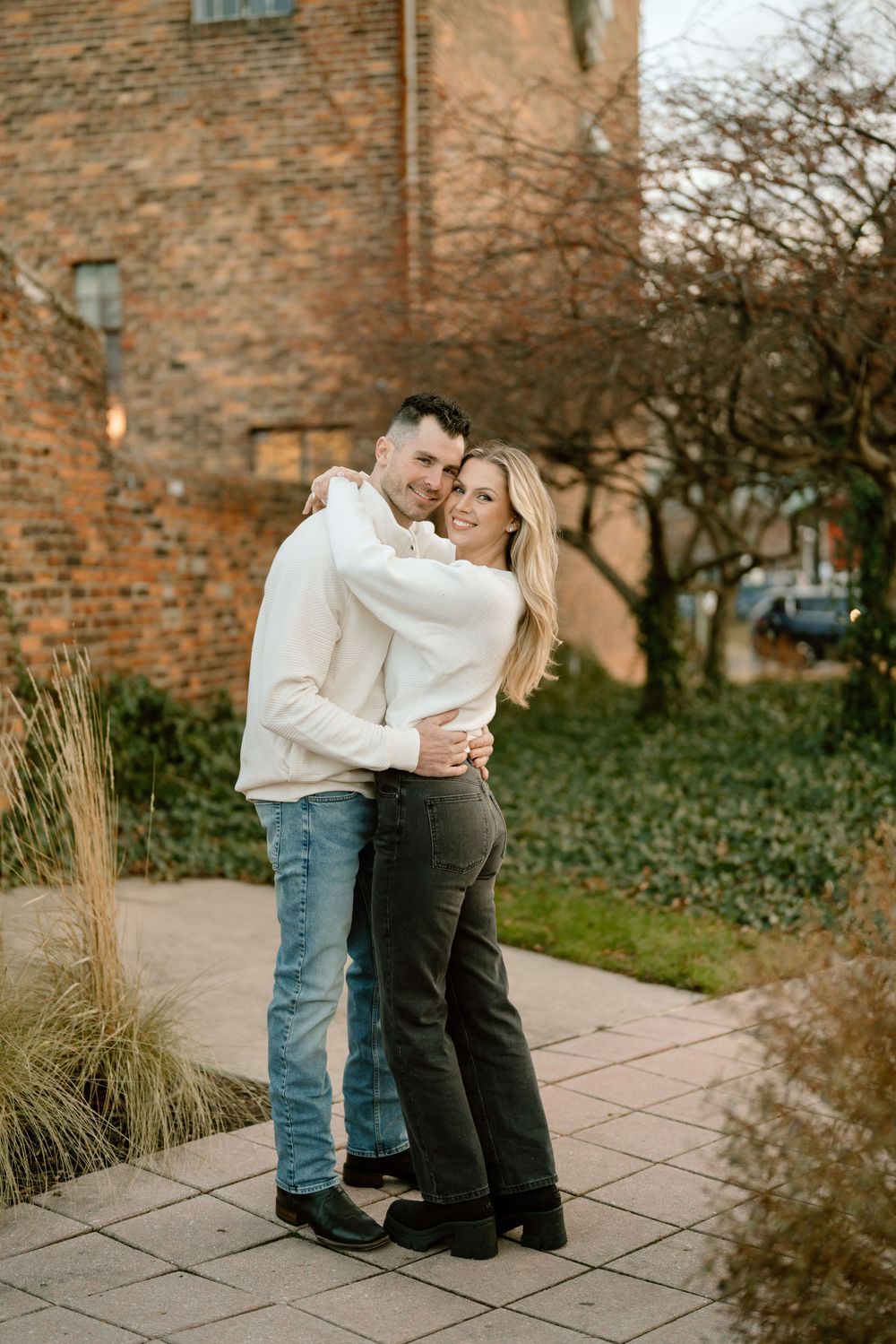 A couple embraces outdoors in casual attire near a brick building and garden path during a romantic moment.