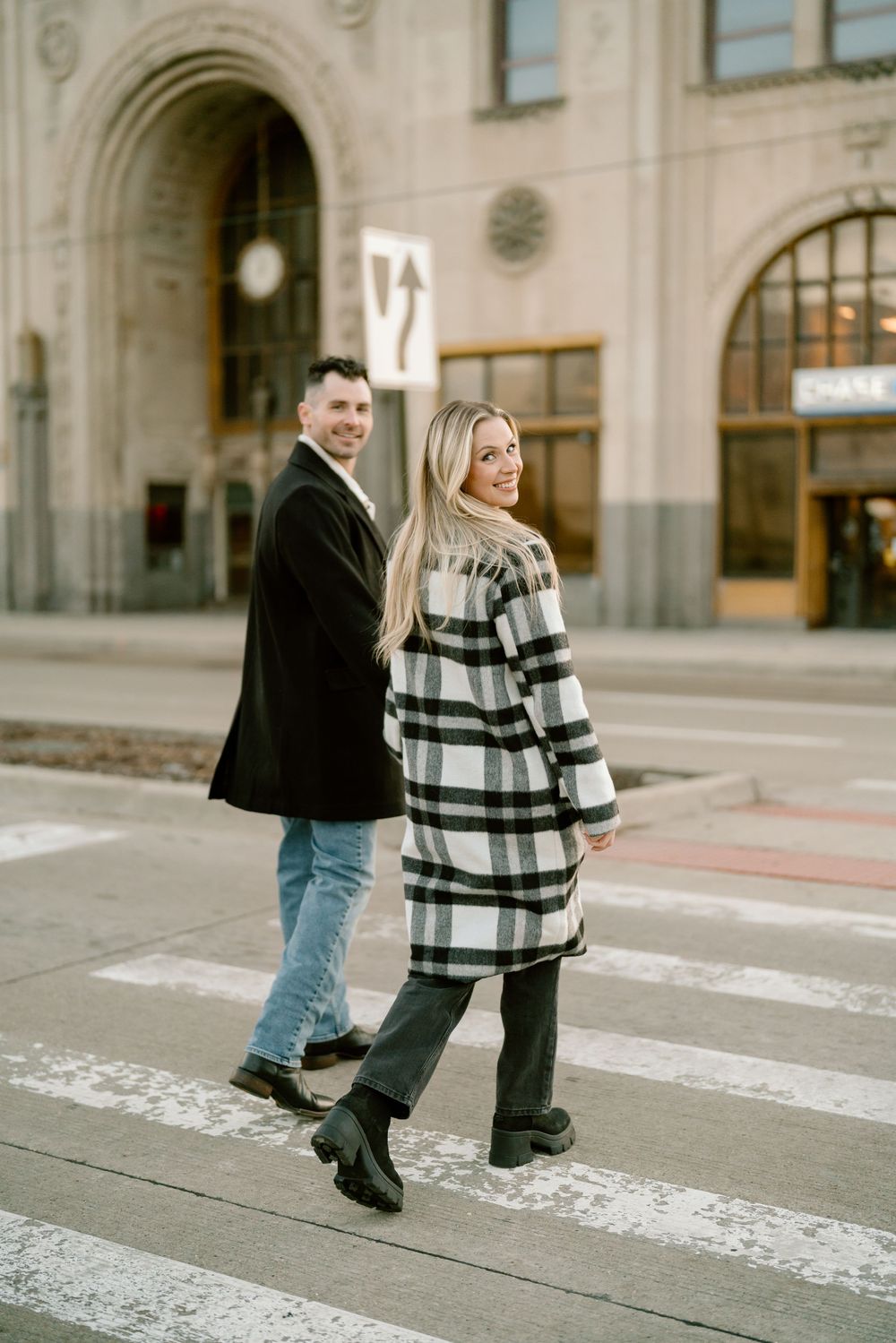 A couple crosses a crosswalk in winter attire, with the woman wearing a striped coat and man in a black peacoat.