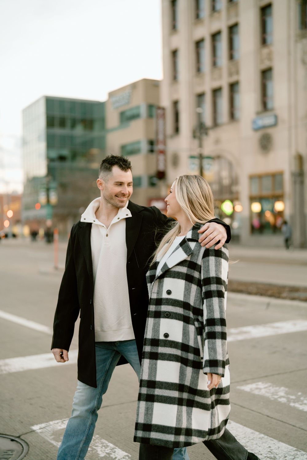 A couple wearing stylish coats walks together on an urban street corner at dusk.
