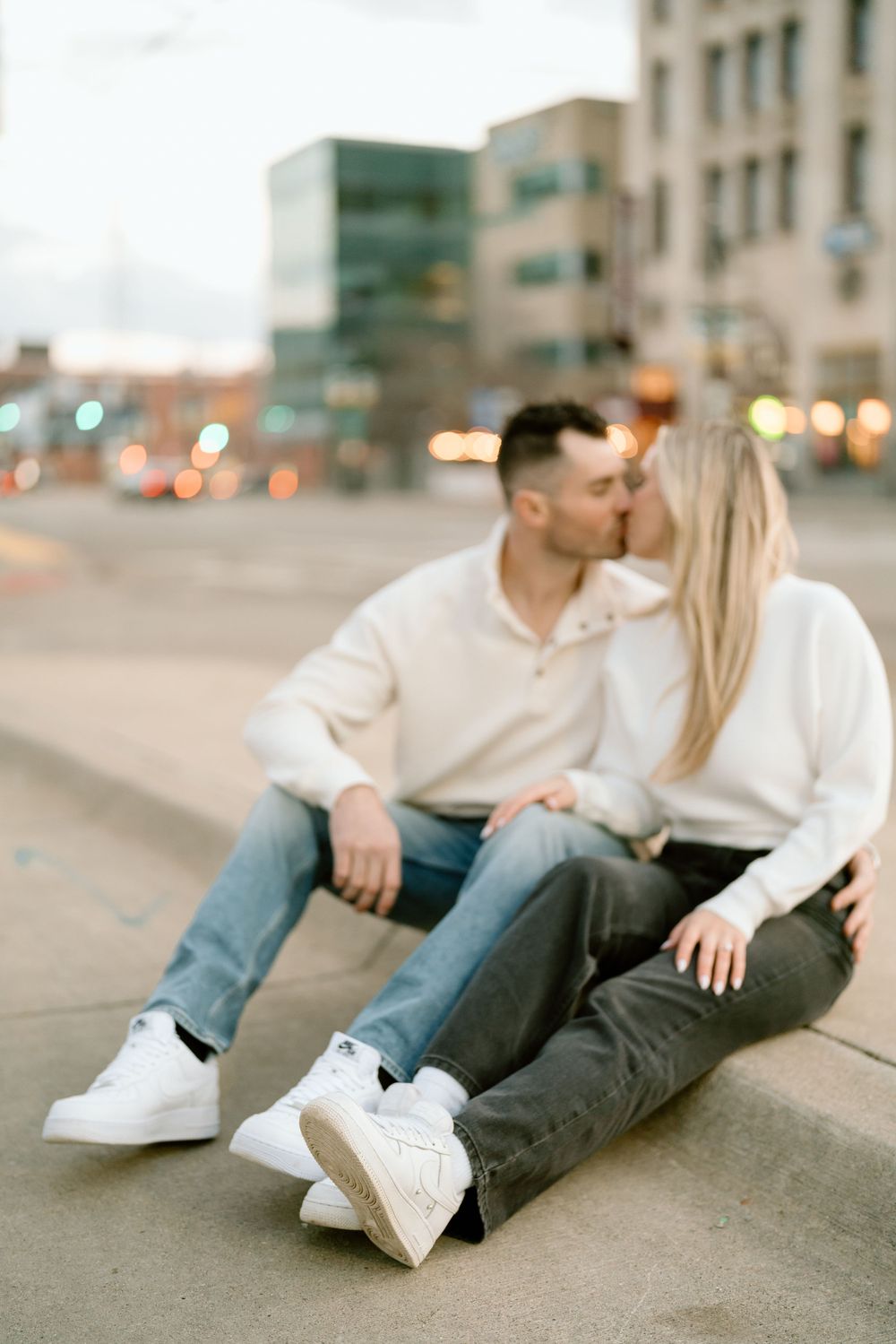 A couple in casual white tops and jeans share a romantic moment sitting on a sidewalk in an urban setting at dusk.