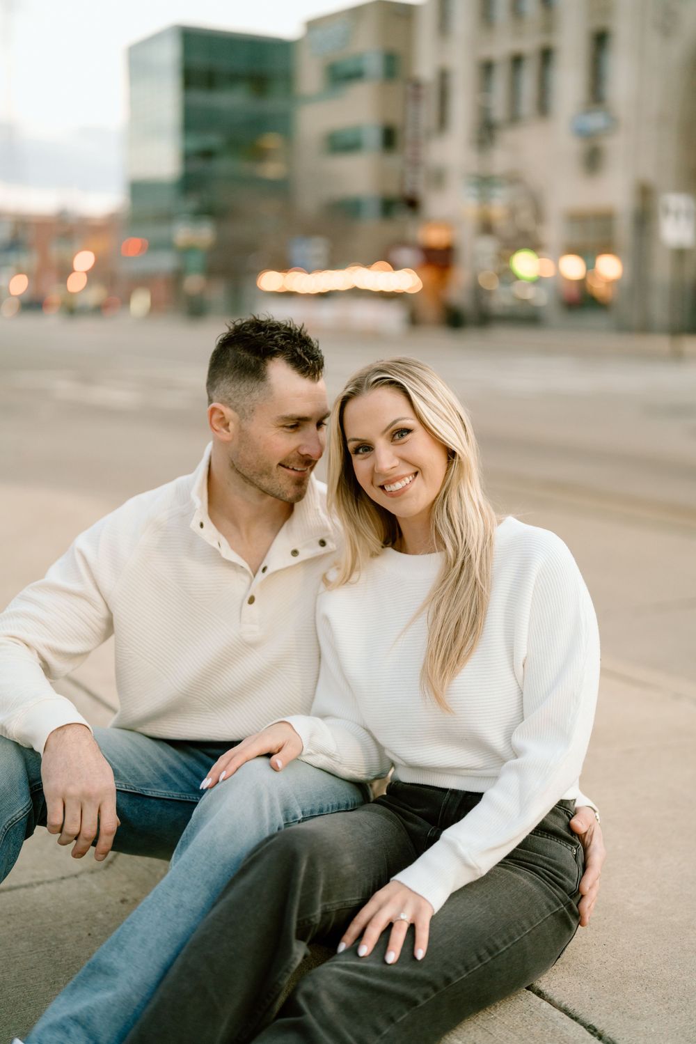 A couple in matching light sweaters sit close together on an urban sidewalk at dusk with blurred city lights behind them.