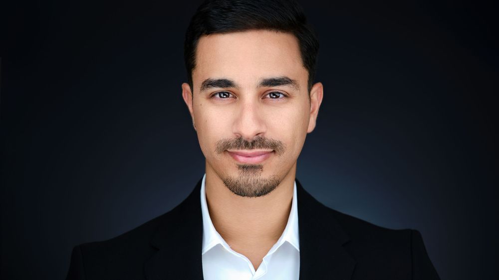 Young man with short dark hair wearing a black blazer and white shirt against a dark background.