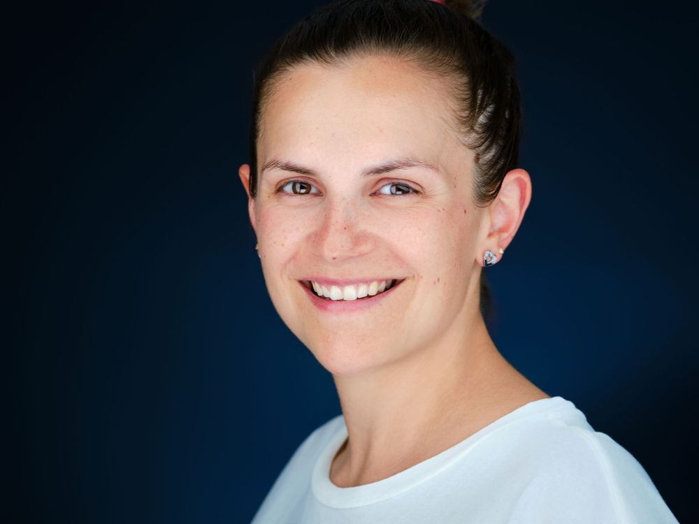 Smiling woman with brown hair pulled back wearing a white top against a dark blue background.