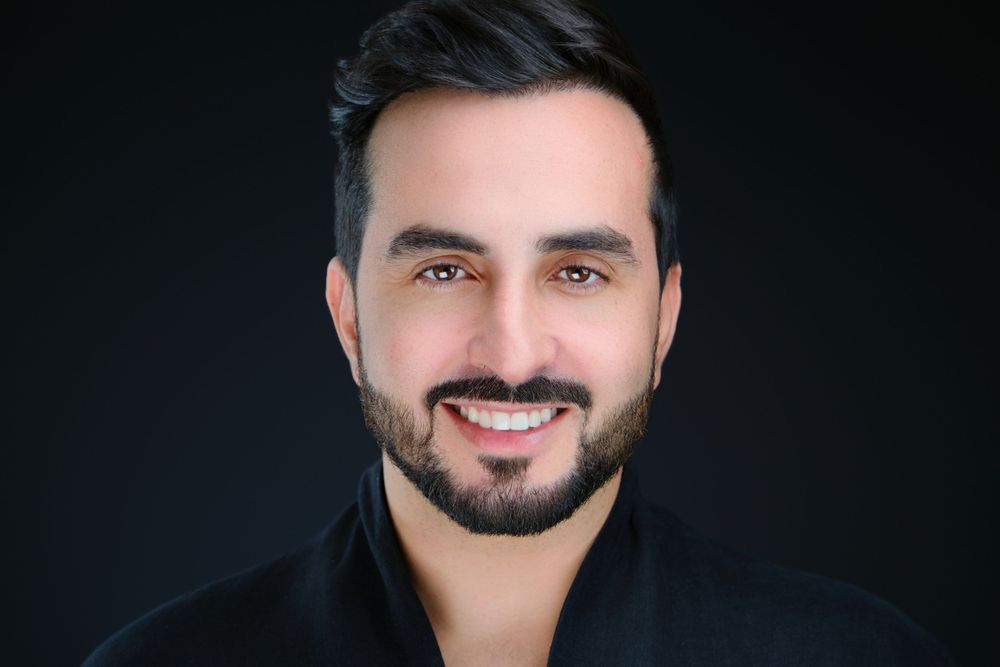 Smiling man with dark beard wearing a black shirt against a dark background.