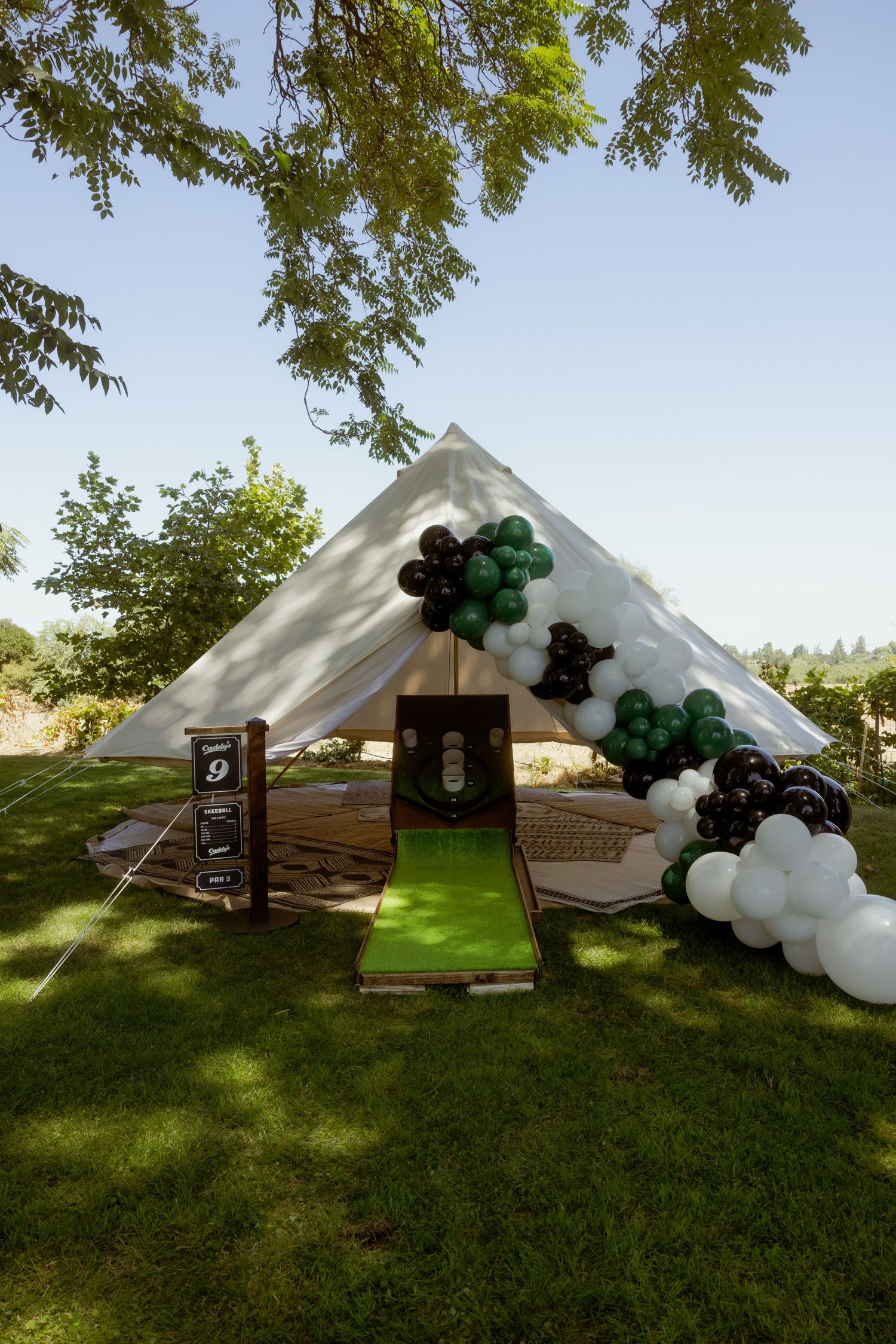 A white tent decorated with black and green balloon garlands and a green carpet entrance in an outdoor garden setting.
