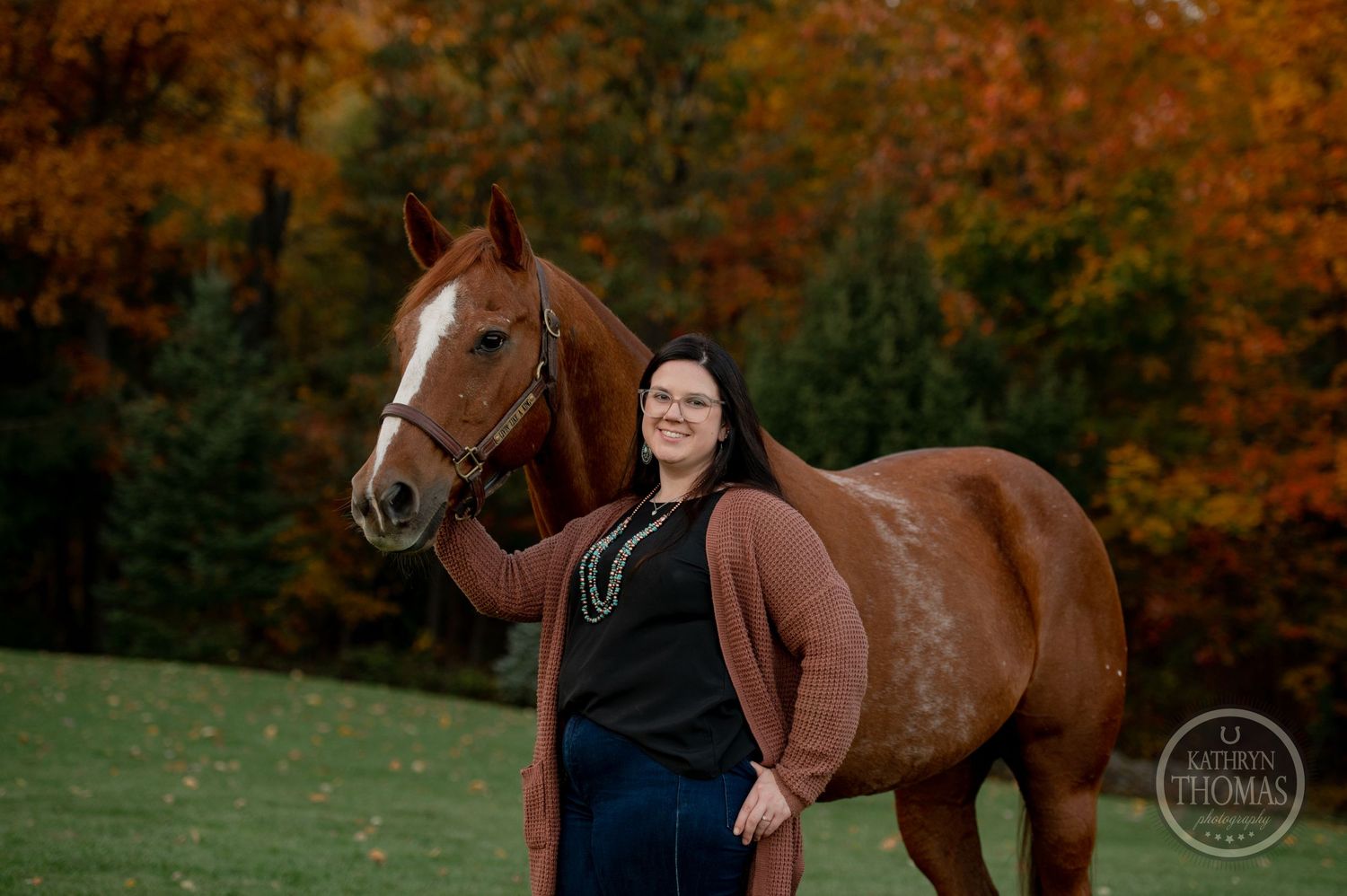 A chestnut horse and person pose together on a grassy slope with autumn trees in background.