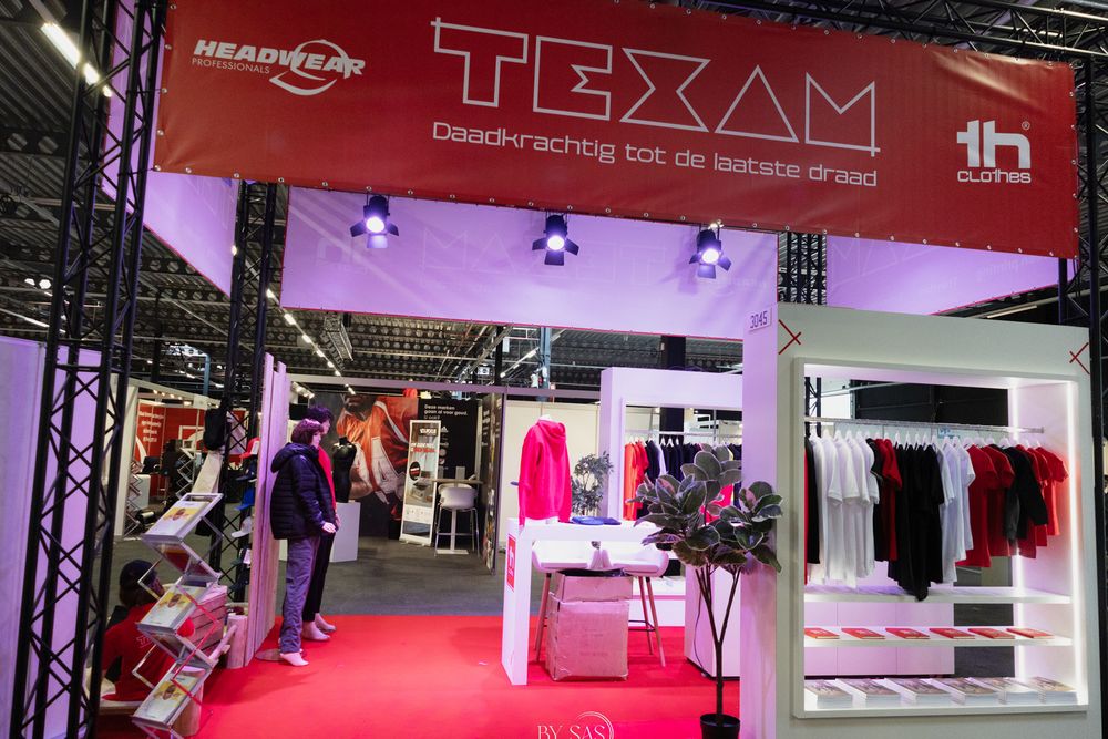 Texam clothing brand trade show booth displaying colorful apparel on racks with red carpet flooring at an indoor exhibition.