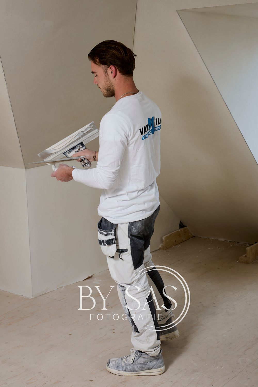 A plasterer in white work clothes holds a plastering tool while working on interior walls of a building under renovation.