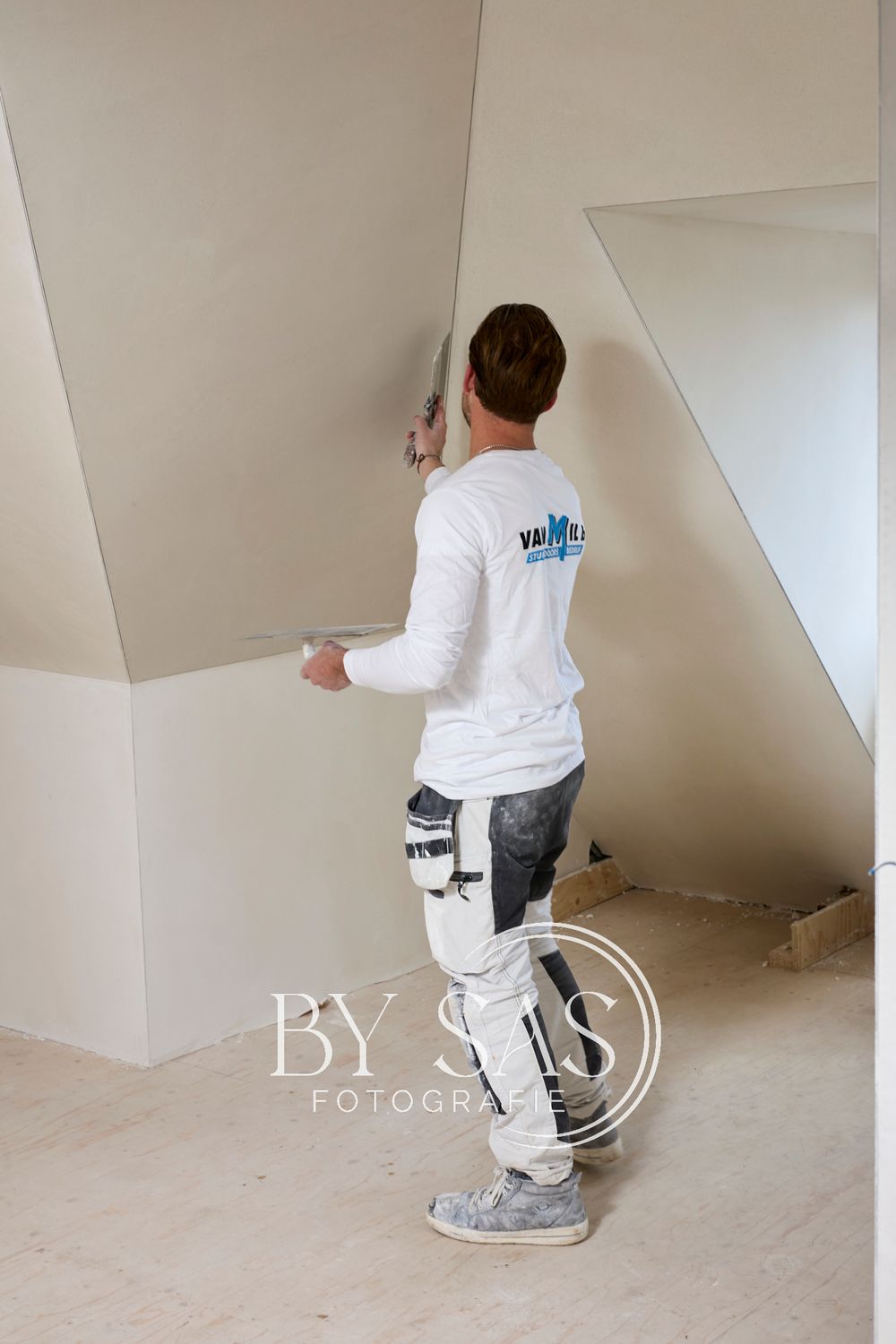 Worker in white uniform applying plaster to a wall in a room under renovation.