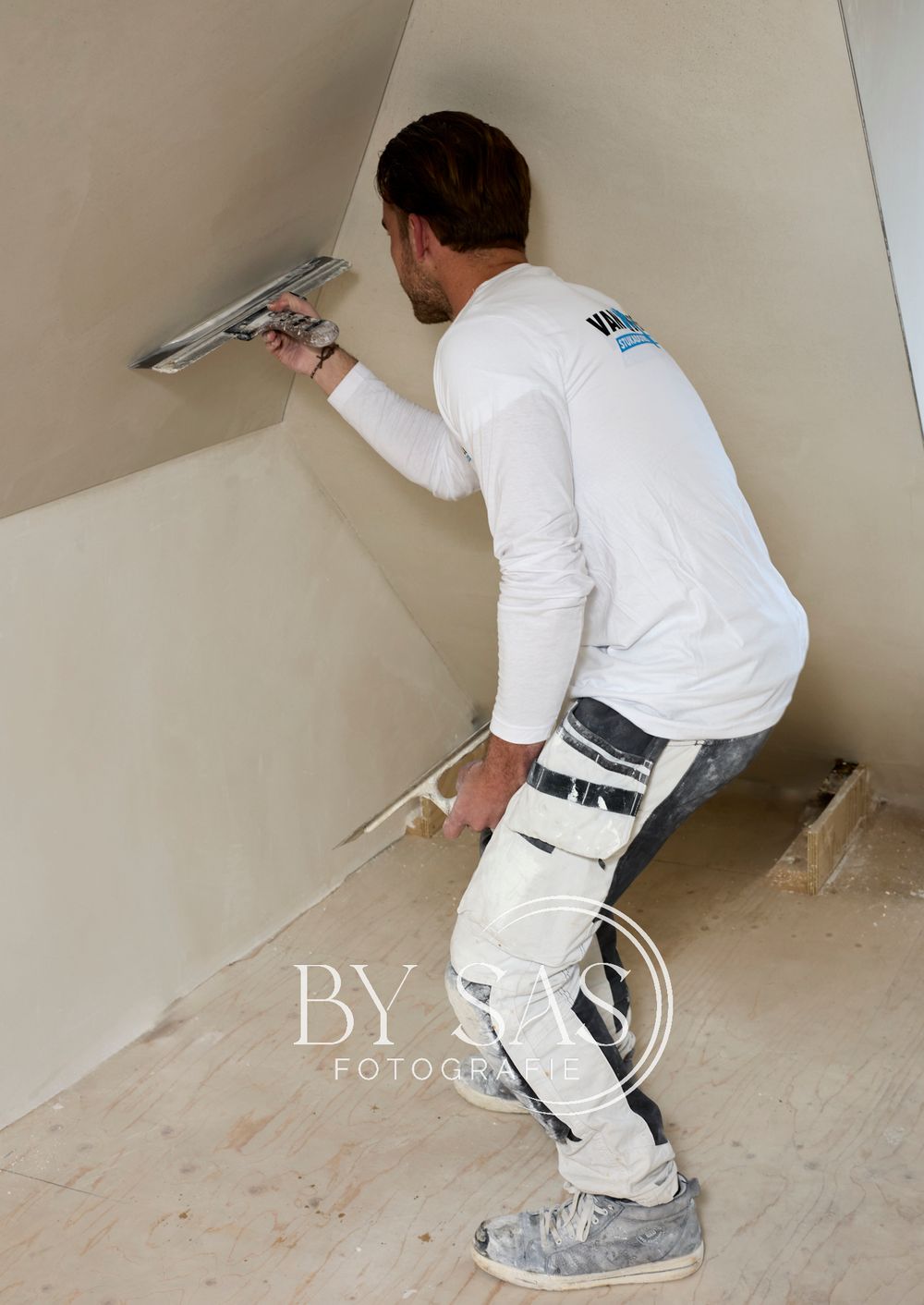 A plasterer wearing a knee brace applies plaster to a wall using a steel trowel during home renovation.