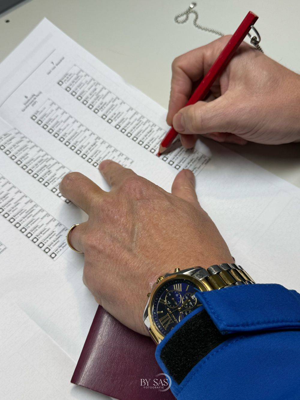 Person filling out a ballot or survey form with a red pencil, wearing a gold watch and blue sleeve.