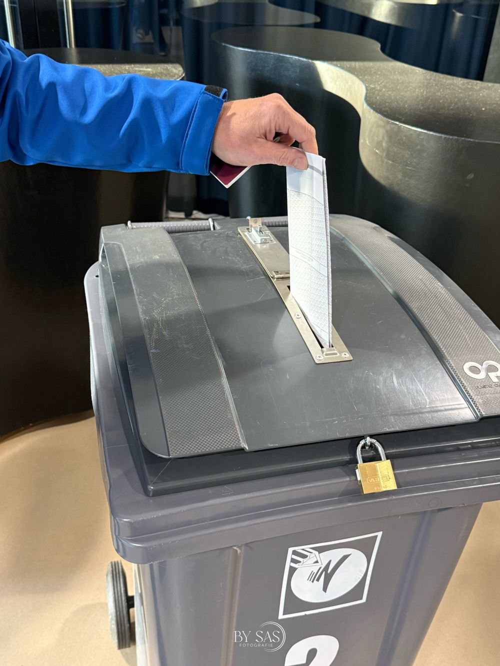 Person in blue jacket inserting a ballot into a secured gray ballot box with padlock during an election.