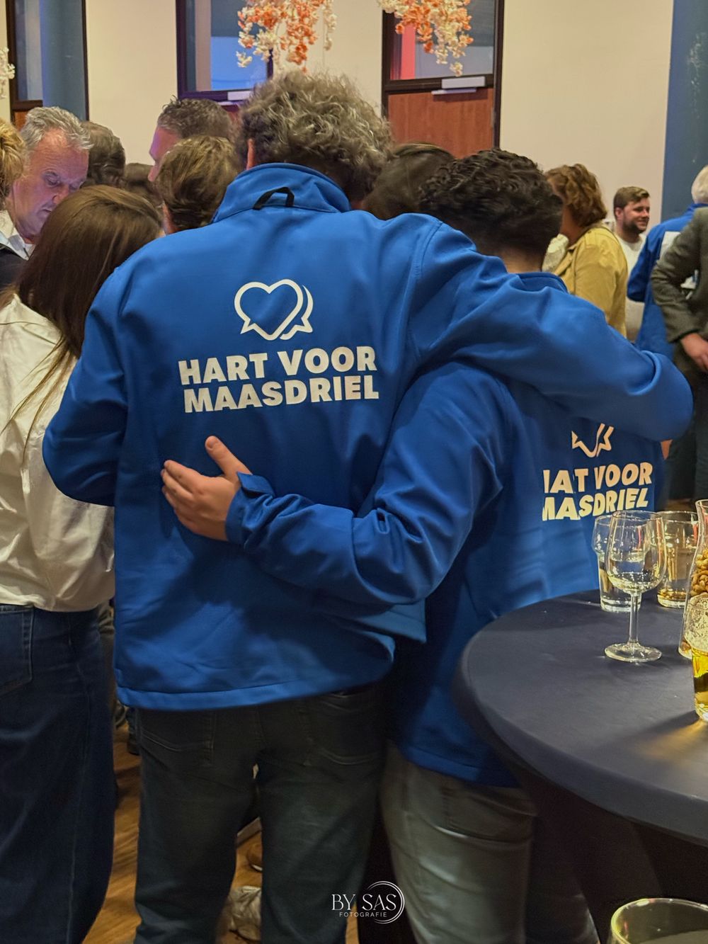 People in blue Hart Voor Maasdriel jackets embracing at a political party gathering event.