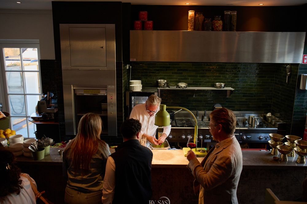 Chef demonstrating cooking techniques to guests at a modern kitchen counter with green tile backsplash.