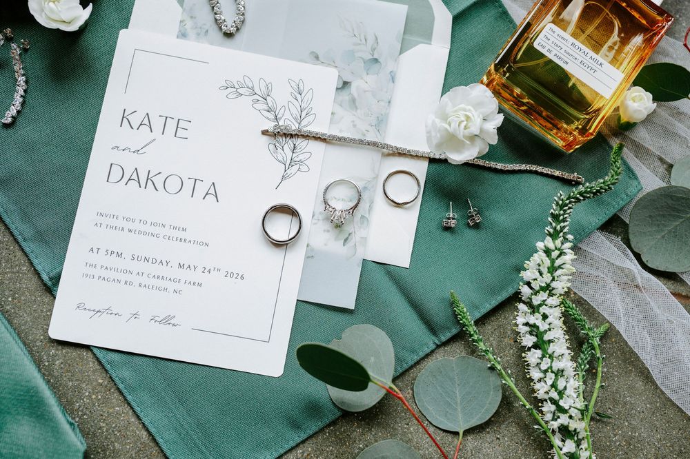 Elegant wedding flat lay with Kate and Dakota invitation, gold rings, pearl jewelry, and green ribbon on teal fabric.