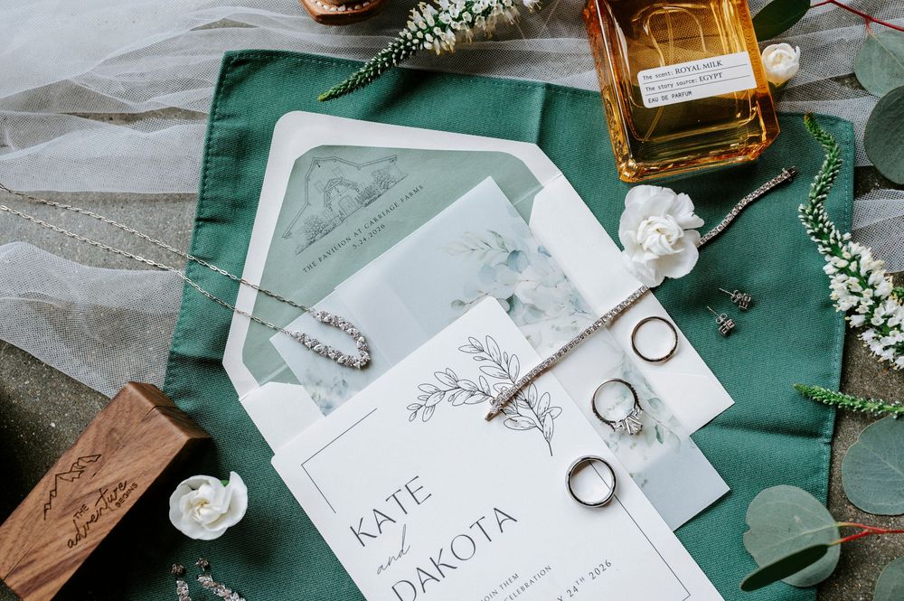 Elegant wedding flat lay with Kate and Dakota invitation suite, rings, perfume, and greenery on emerald green fabric.