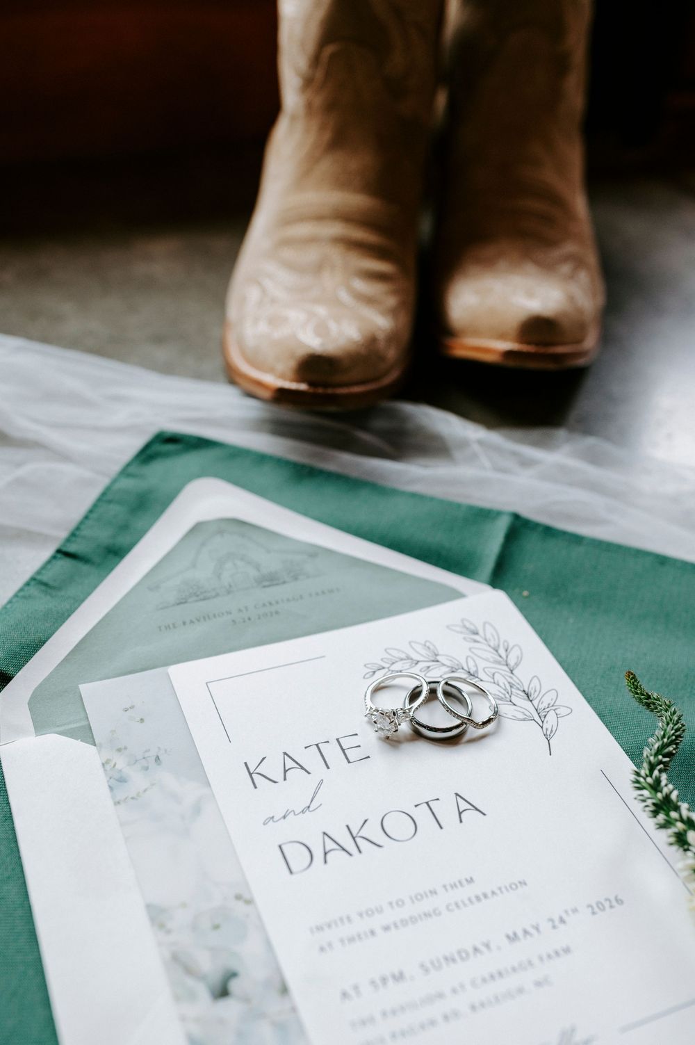 Kate and Dakota wedding invitation suite with diamond rings and cowboy boots on green fabric for rustic celebration.