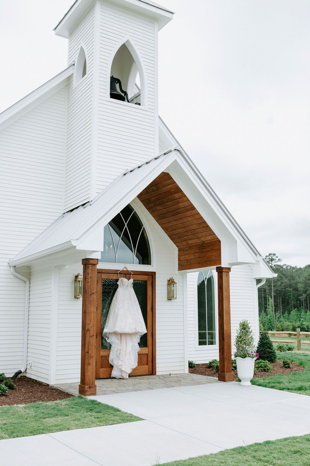 White chapel wedding venue with wooden entrance doors displaying an elegant bridal gown hanging outside.