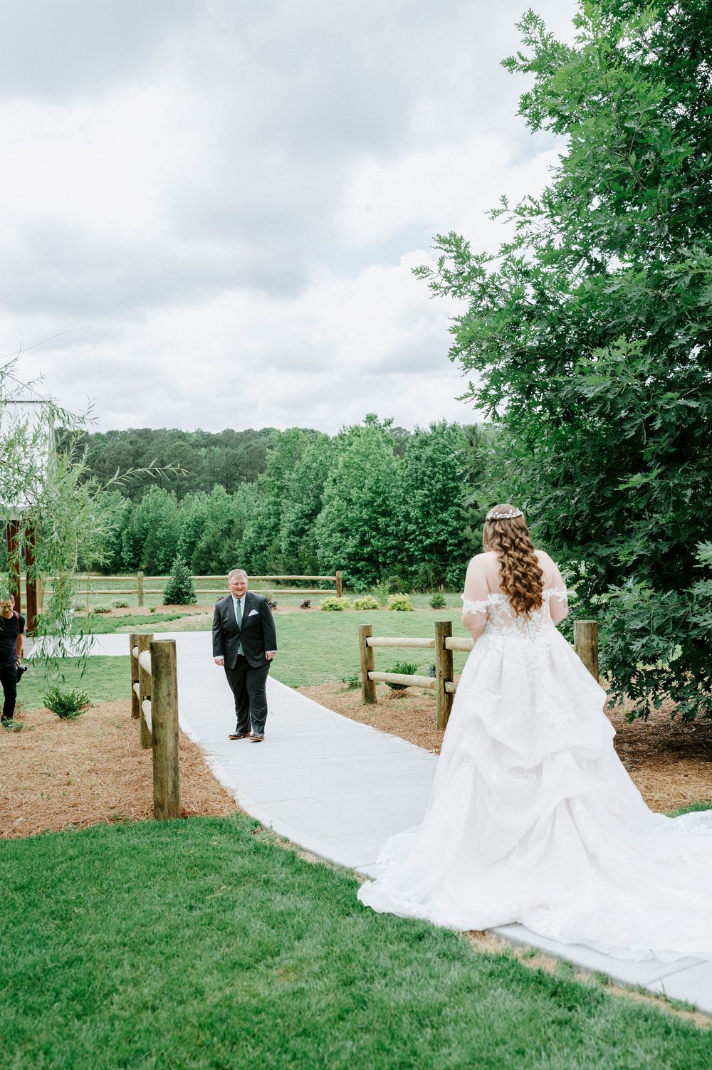 Bride in white gown with long train stands on garden path as man in suit approaches during outdoor wedding ceremony.
