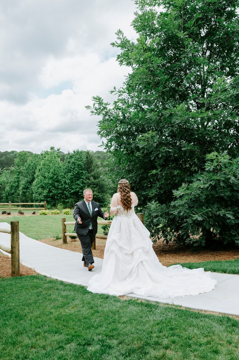 Bride and groom walking hand-in-hand along a white pathway surrounded by lush green trees.