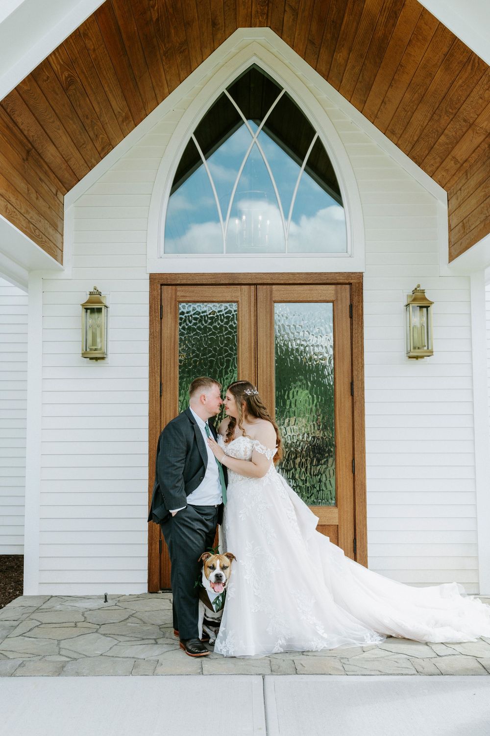 Bride and groom share a kiss outside a white chapel with their dog in a green bandana on their wedding day.