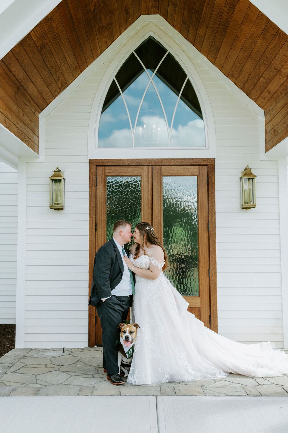Bride and groom share a romantic moment with their dog outside a white chapel with wooden doors and arched window.