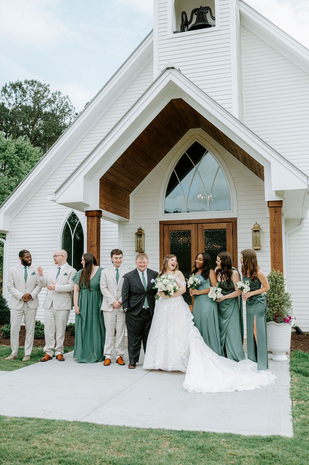 Wedding party in sage green attire poses outside a white chapel with arched windows and wooden doors.