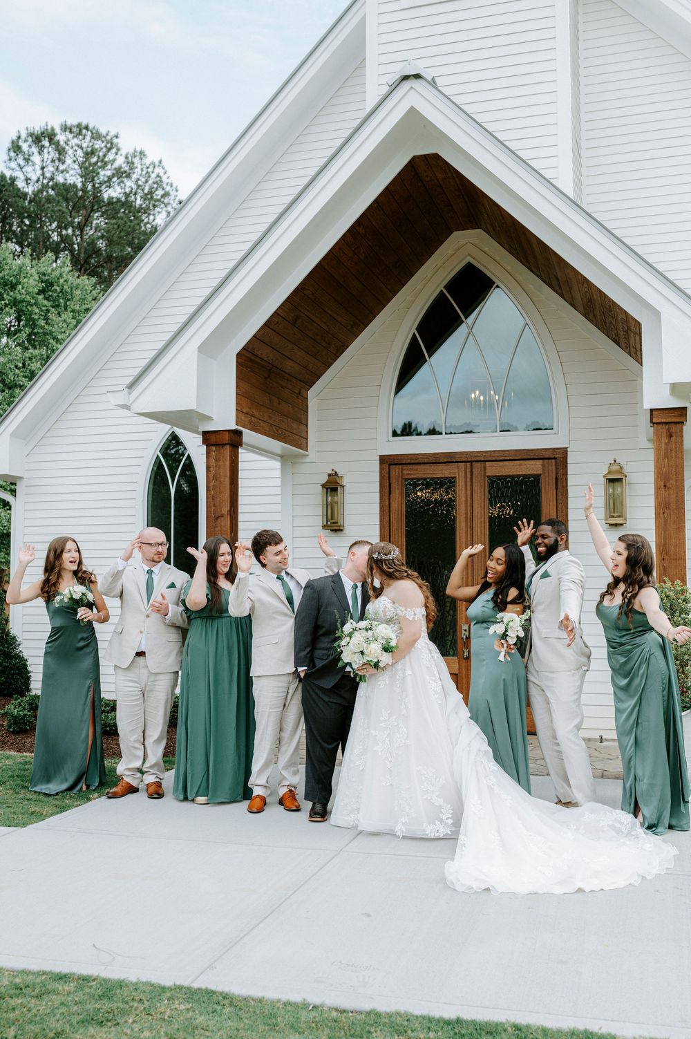 Wedding party in sage green dresses and gray suits celebrating outside a white chapel venue.