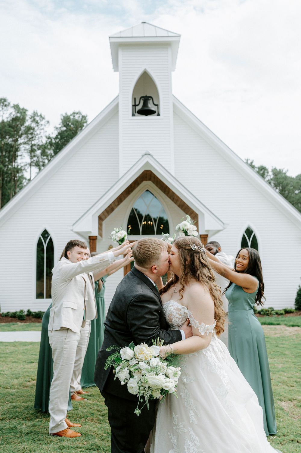 Bride and groom kiss outside a white country chapel surrounded by wedding party in sage green and white florals.