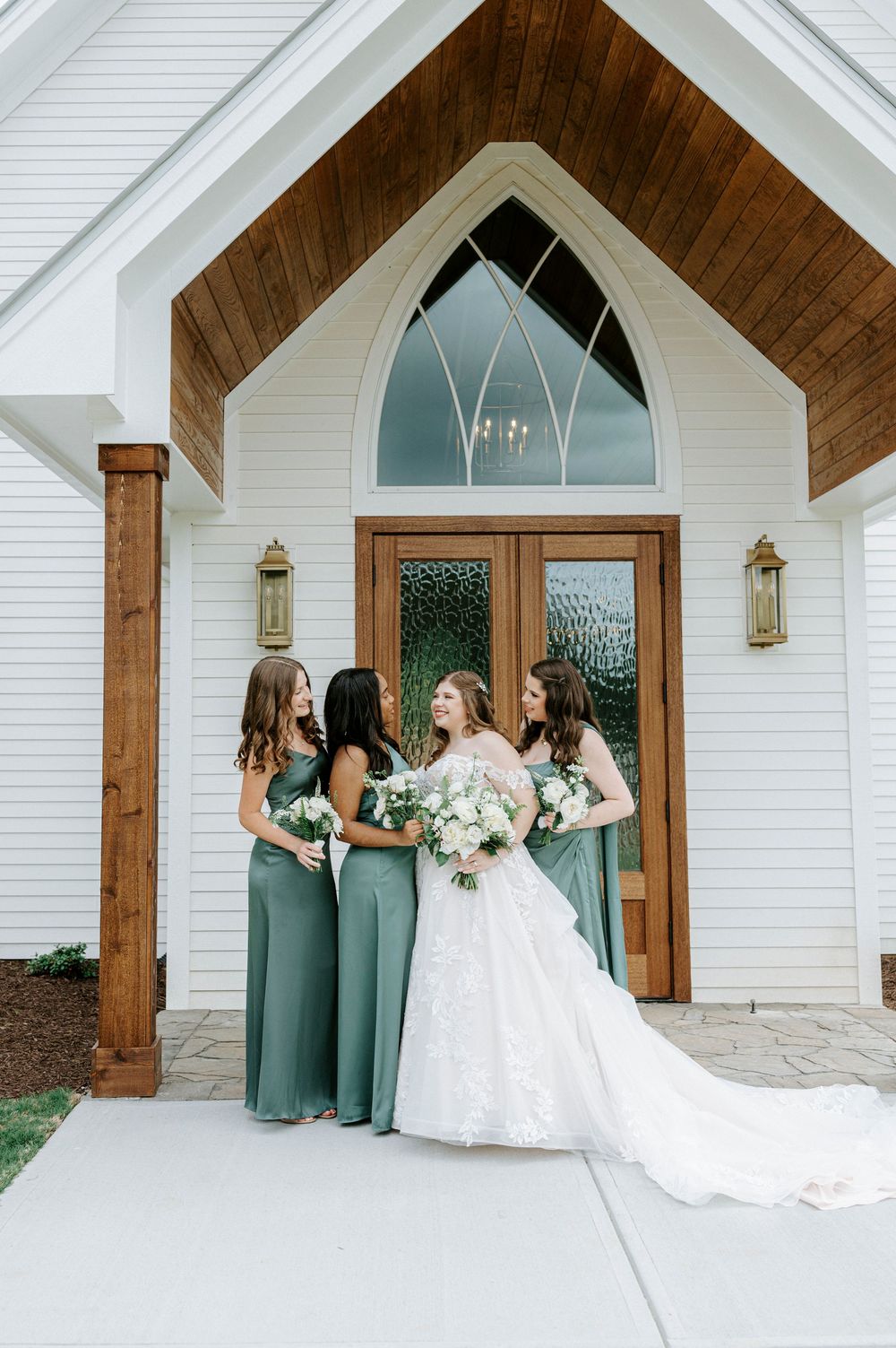 Bride in lace gown holds white bouquet with three bridesmaids in sage green dresses outside a white chapel.