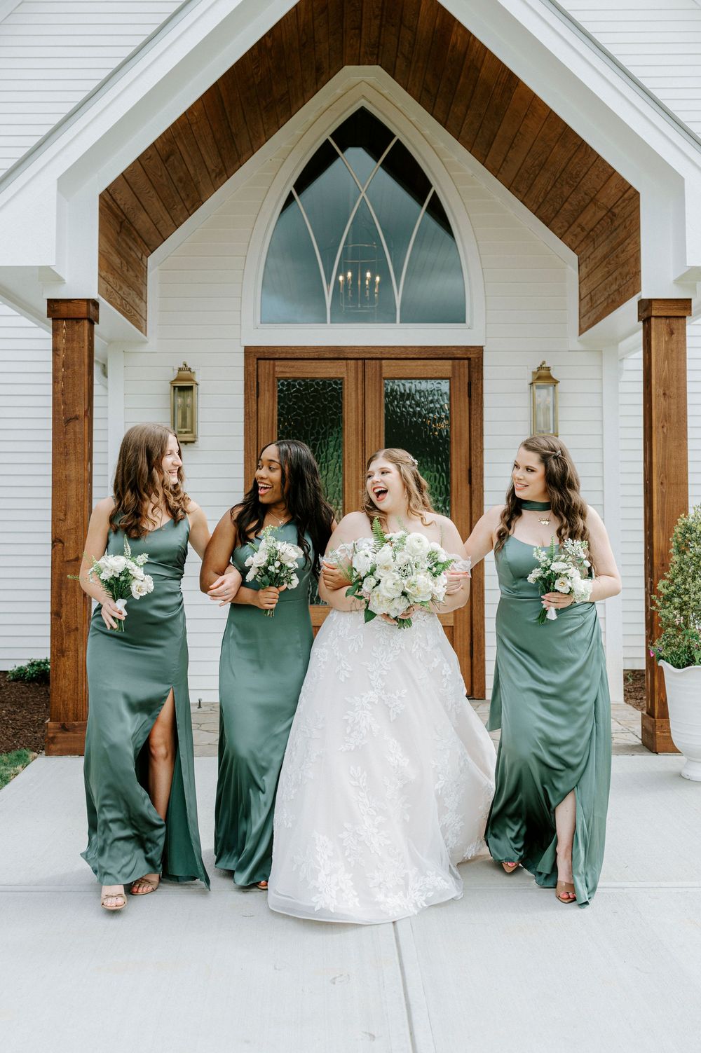 Bride laughing joyfully with three bridesmaids in sage green satin dresses holding white bouquets outside a chapel.