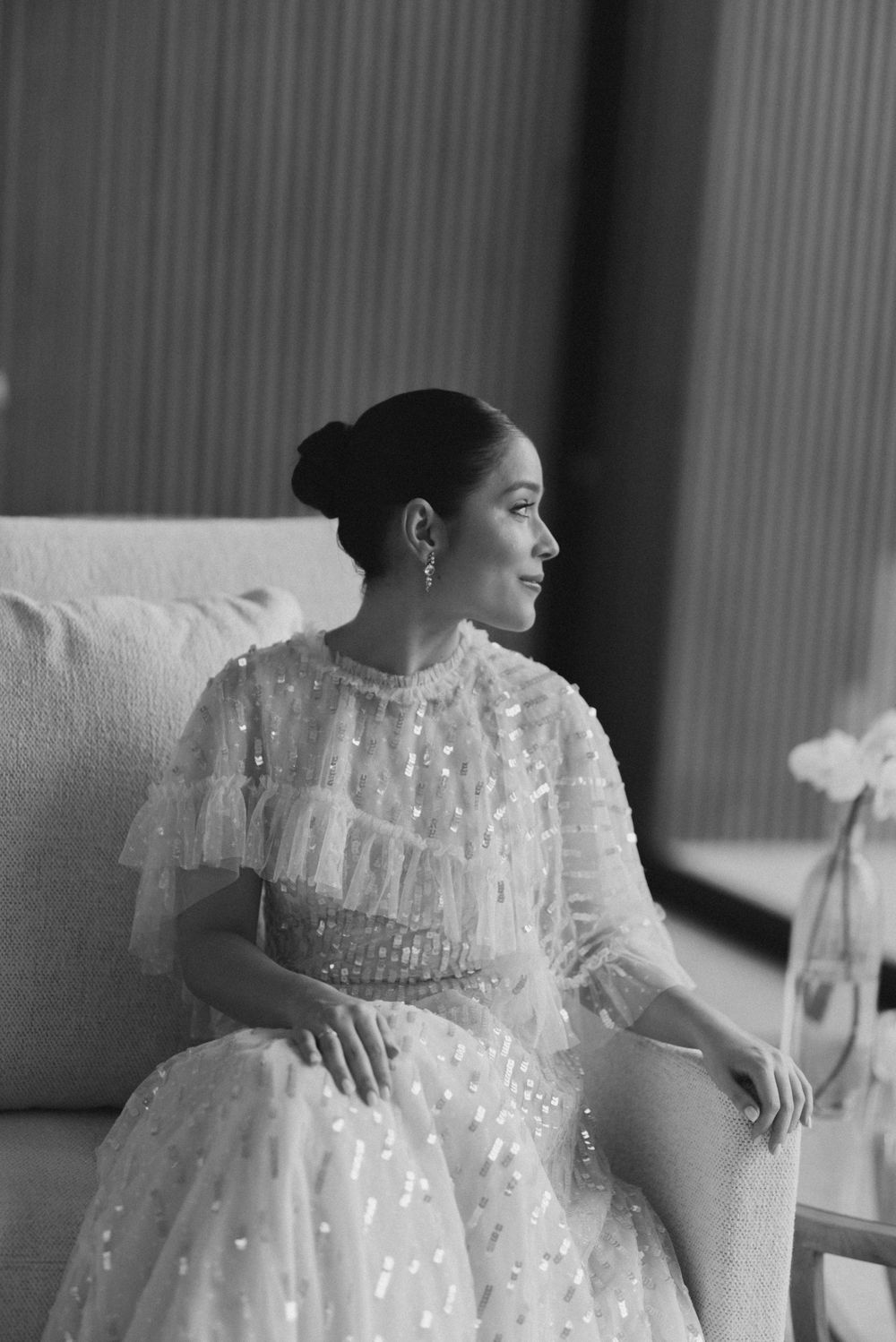 Black and white portrait of someone in an elegant white lace dress with sparkles sitting on a couch by a window.