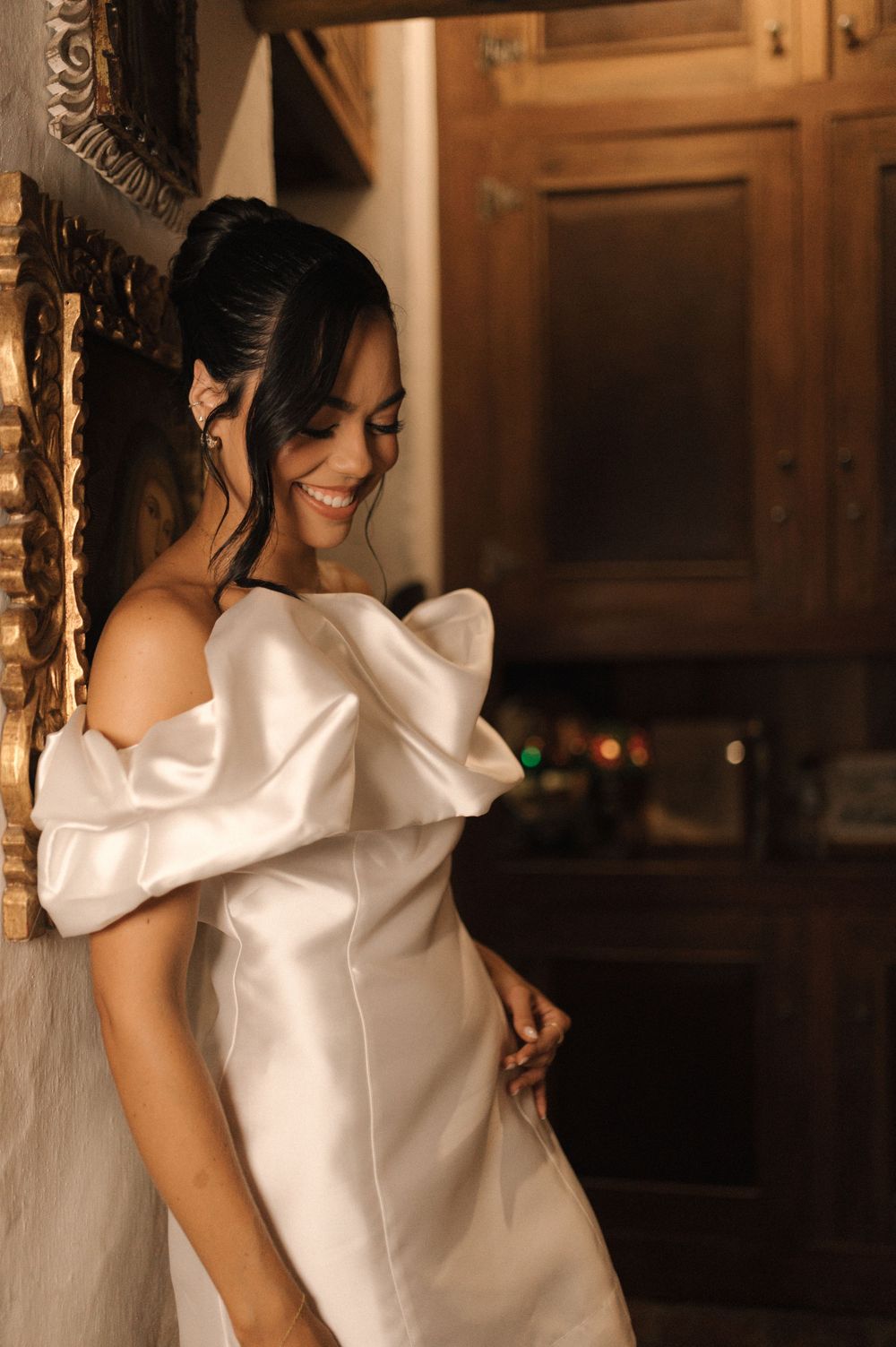 Smiling bride in elegant off-shoulder white satin wedding gown posing in a warmly lit rustic interior.