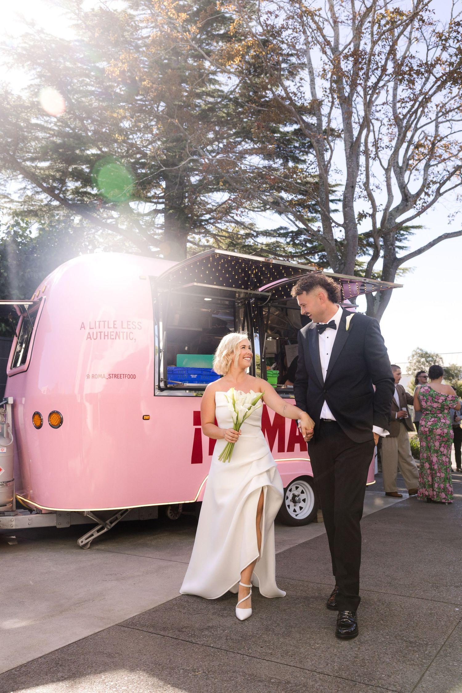 Bride and groom walking hand in hand beside a pink vintage trailer at their outdoor wedding reception.
