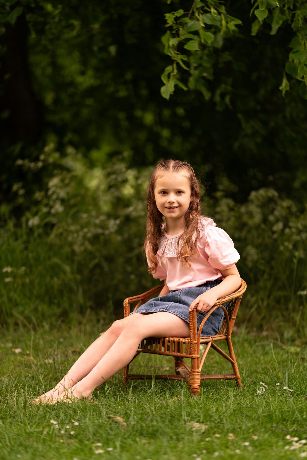 Smiling little girl in pink top and denim skirt posing on wicker chair on green grass in garden during childhood photoshoot in Lincolnshire.