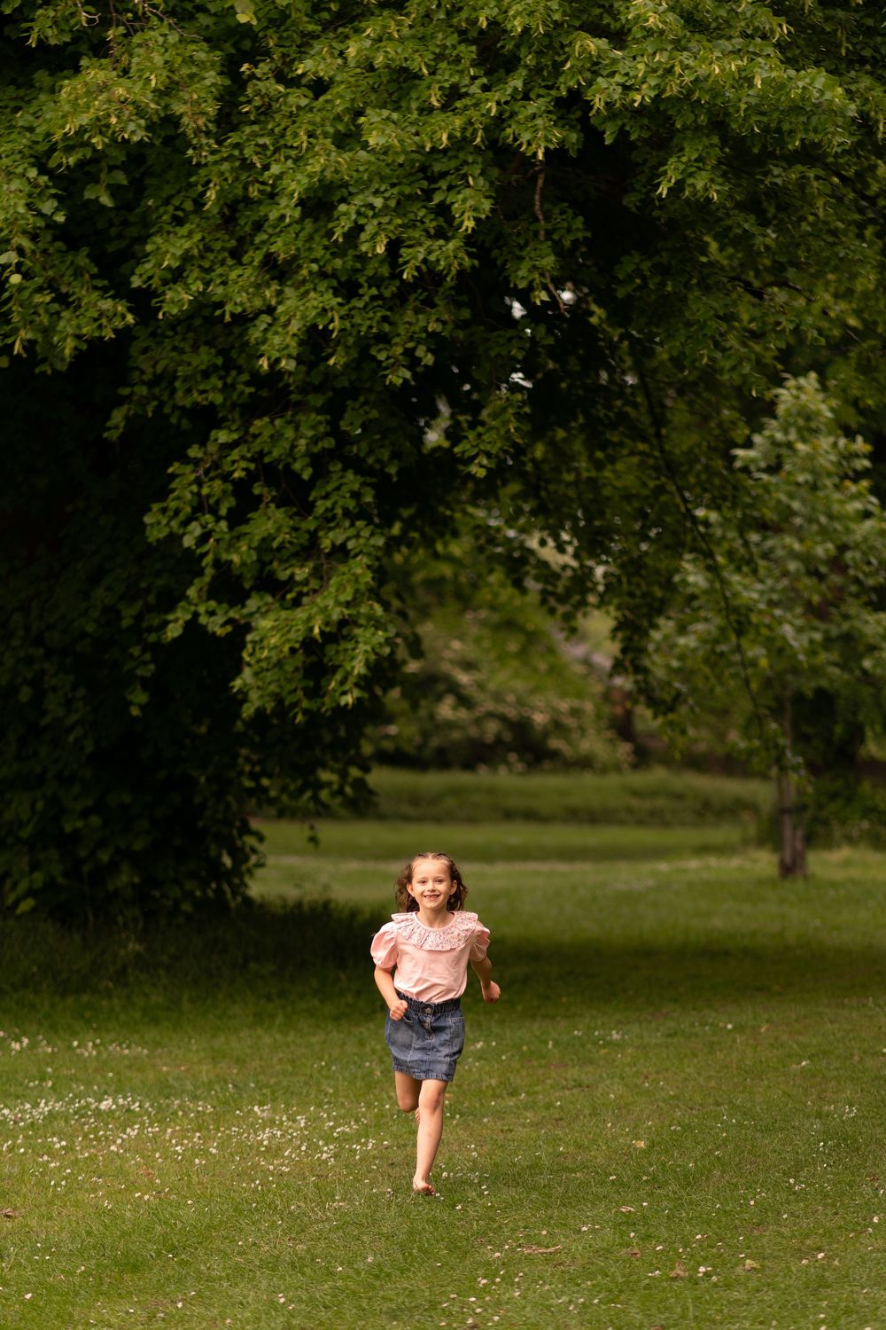 Happy young girl in pink blouse and denim skirt running barefoot across lush green park lawn during childhood photoshoot in Lincolnshire.