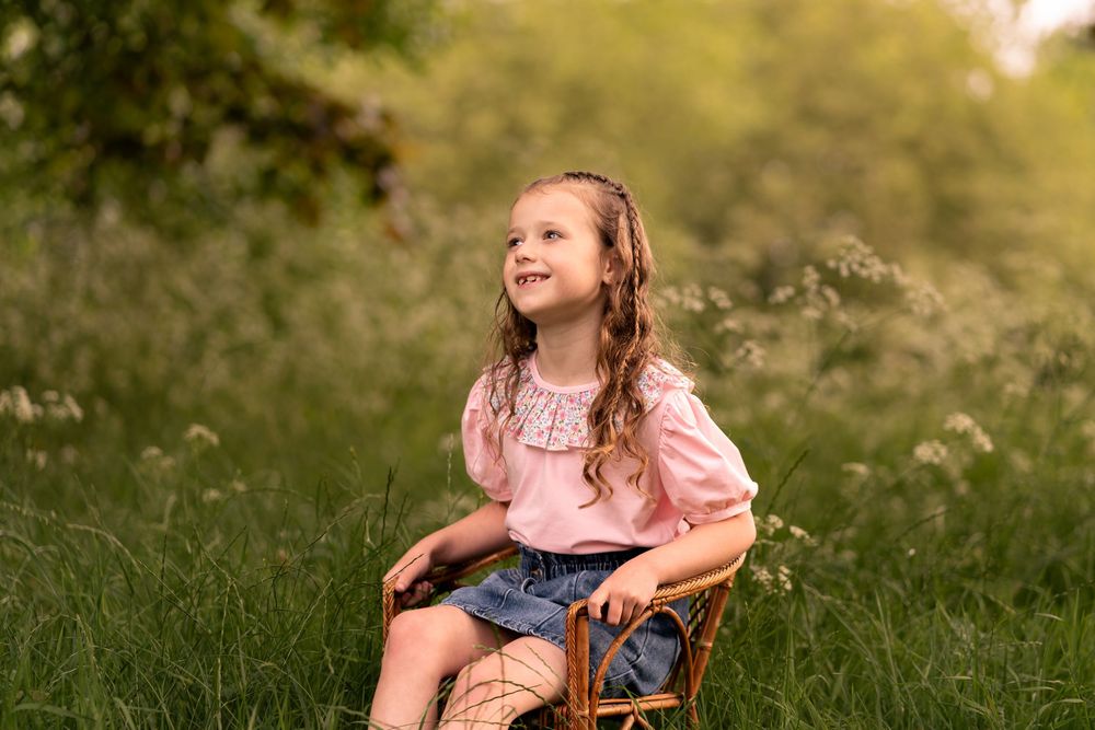 Young girl sitting on wicker chair in sunny meadow with wildflowers during childhood photoshoot in Lincolnshire.