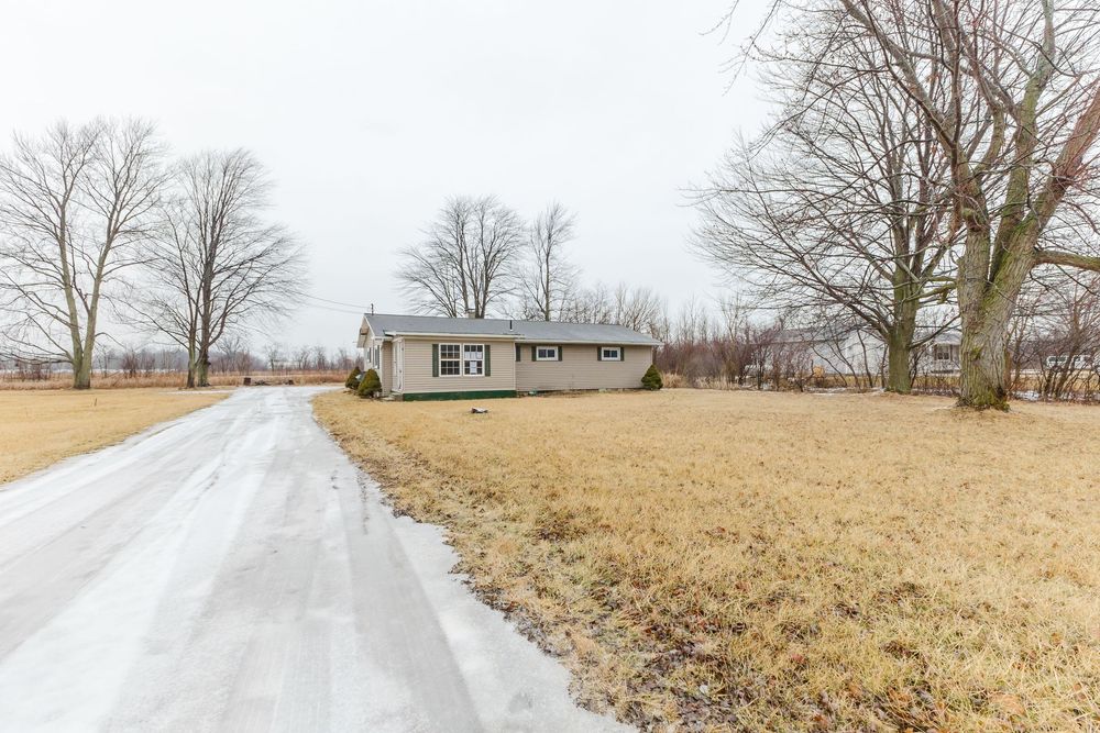 Rural ranch-style home with long driveway, dormant grass lawn, and bare trees under overcast winter sky.