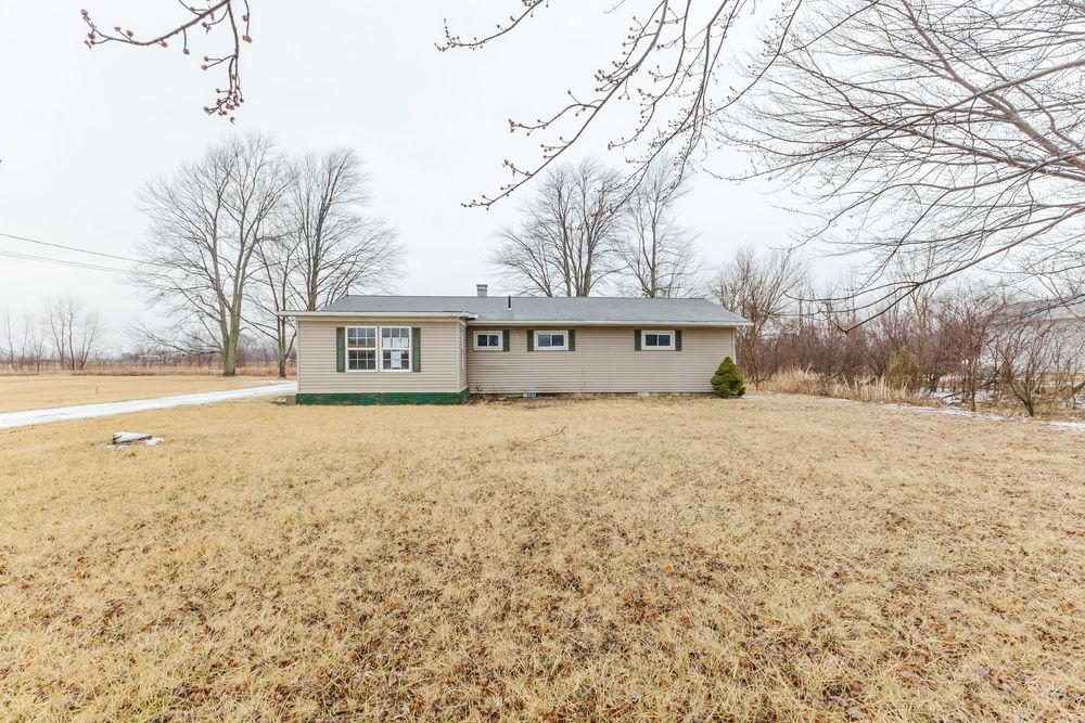 Ranch-style brick home with green shutters sits on a large dormant lawn surrounded by bare winter trees under overcast skies.