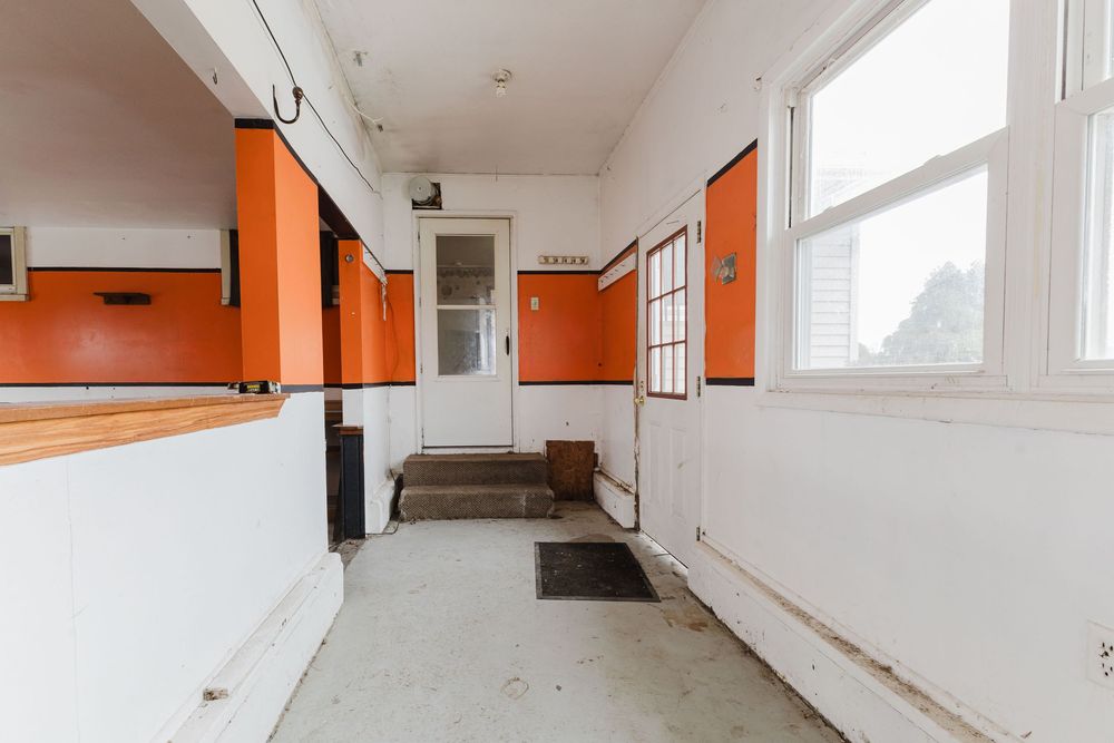 Narrow hallway with bold orange and white walls, concrete floor, and a white door at the end letting in natural light.