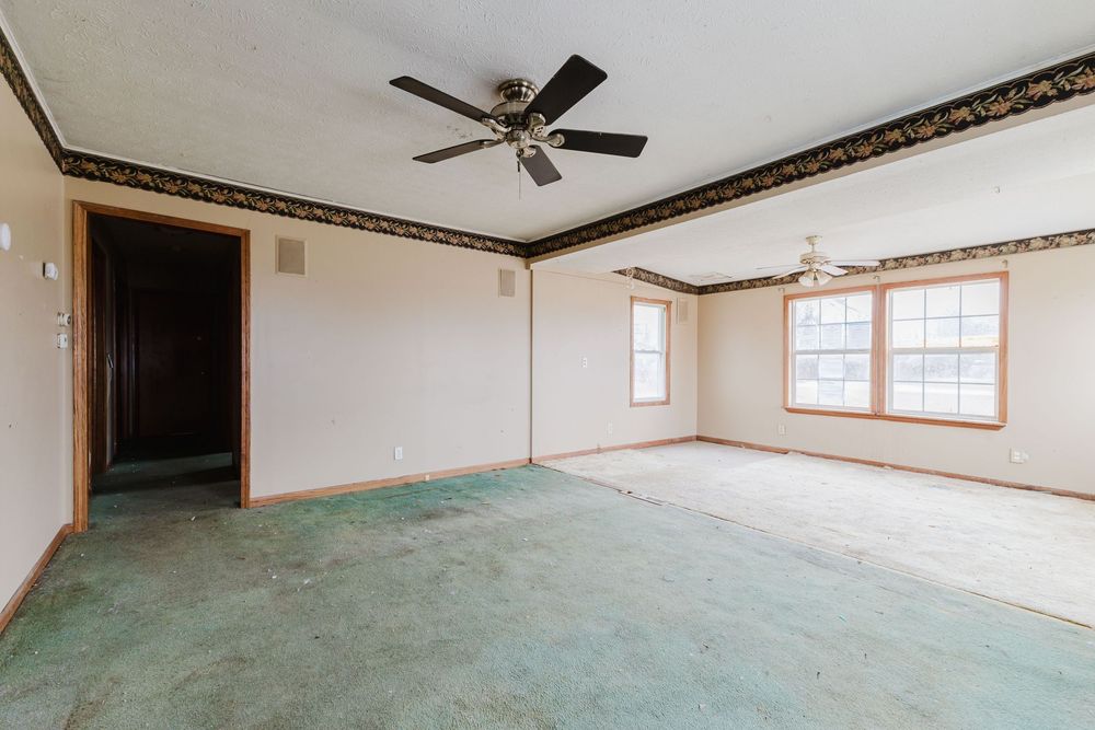 Spacious empty living room with green carpet, ceiling fan, crown molding, decorative border, and natural light from windows.