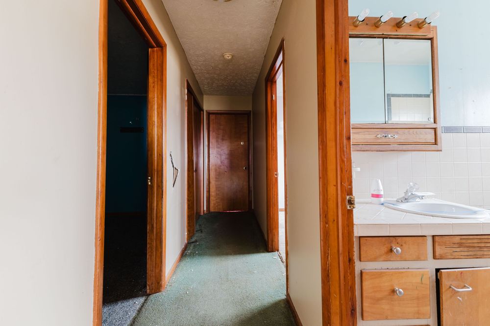 Narrow hallway with green carpet, wood-trimmed doors, and a bathroom vanity with sink and wooden cabinets on the right.
