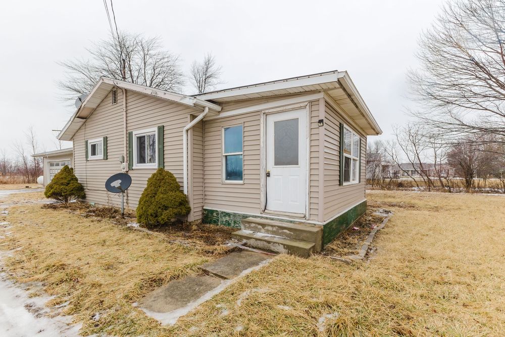 Small ranch-style home with vinyl siding, white entry door, evergreen shrubs, and dormant lawn on a cloudy winter day.