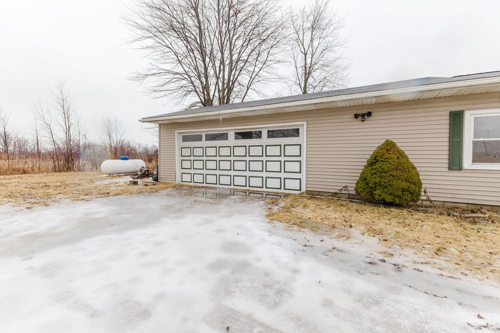 Beige ranch home with white panel garage door, snowy driveway, bare tree, and green shrub in winter.