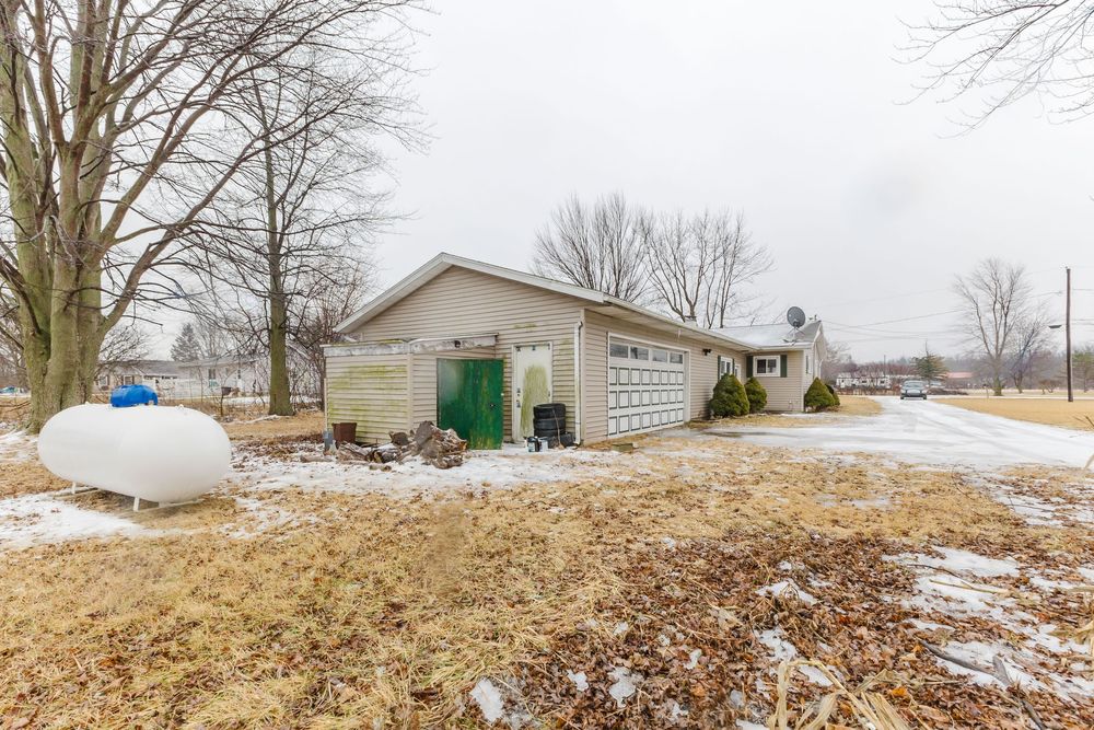 Ranch-style home with attached garage, propane tank, and snow-dusted yard surrounded by bare winter trees.