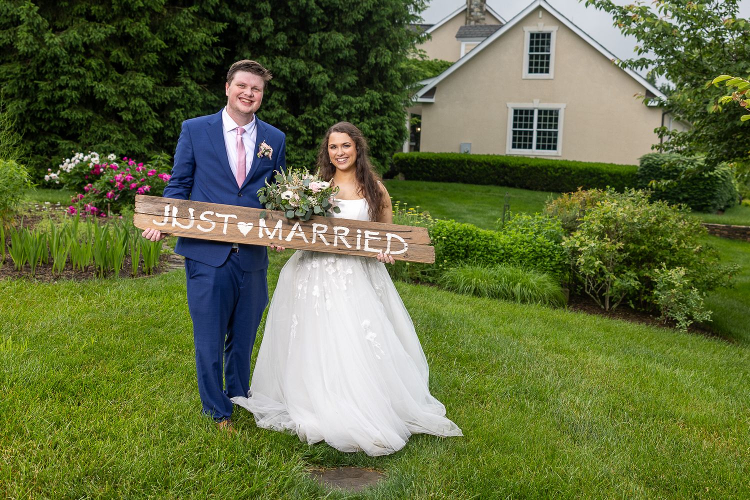 Newlywed couple holding a Just Married wooden sign on green lawn outside a white wedding venue.